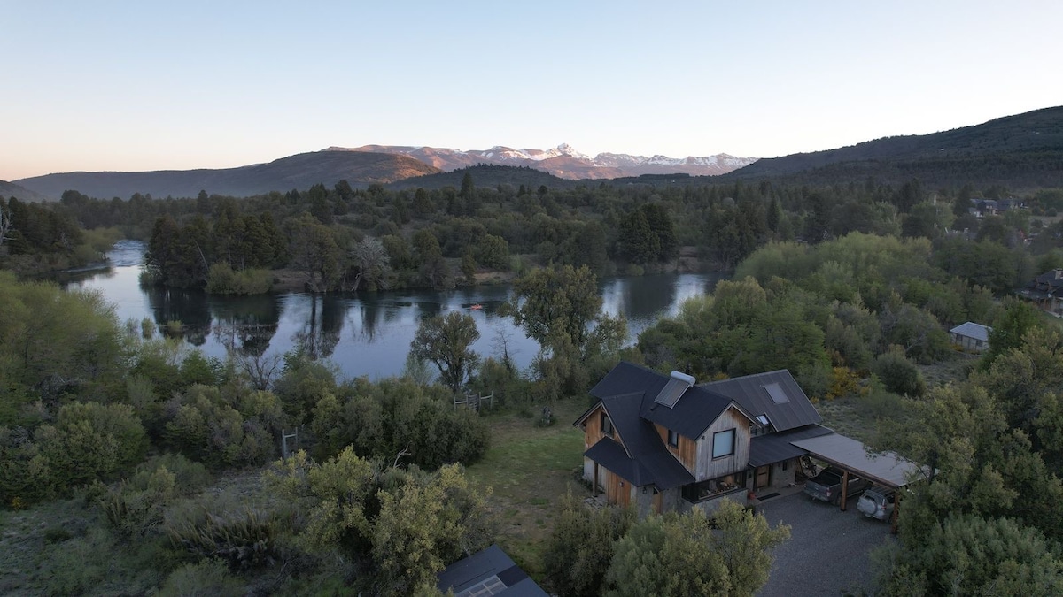 An aerial view showcases a spacious house set amidst lush greenery. The property gently slopes towards the river Quilquihue, with mountains visible in the background under a soft blue sky. Natural light highlights the surrounding trees and the tranquil water.