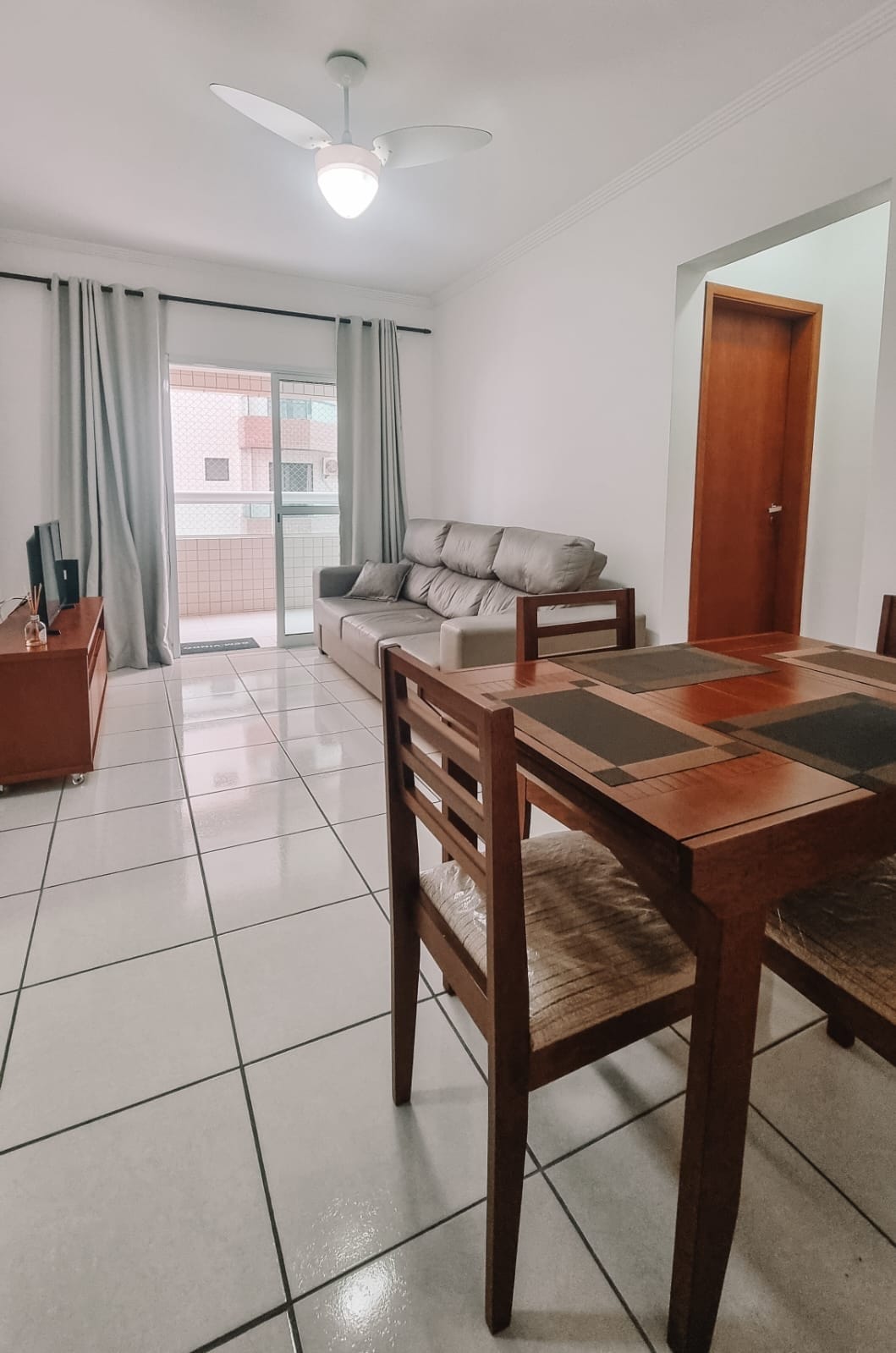 A comfortable living space is presented, featuring a light-colored sofa and a small dining table with four chairs. Large glass doors allow natural light and access to the balcony, while tiled floors enhance the room's spacious feel.