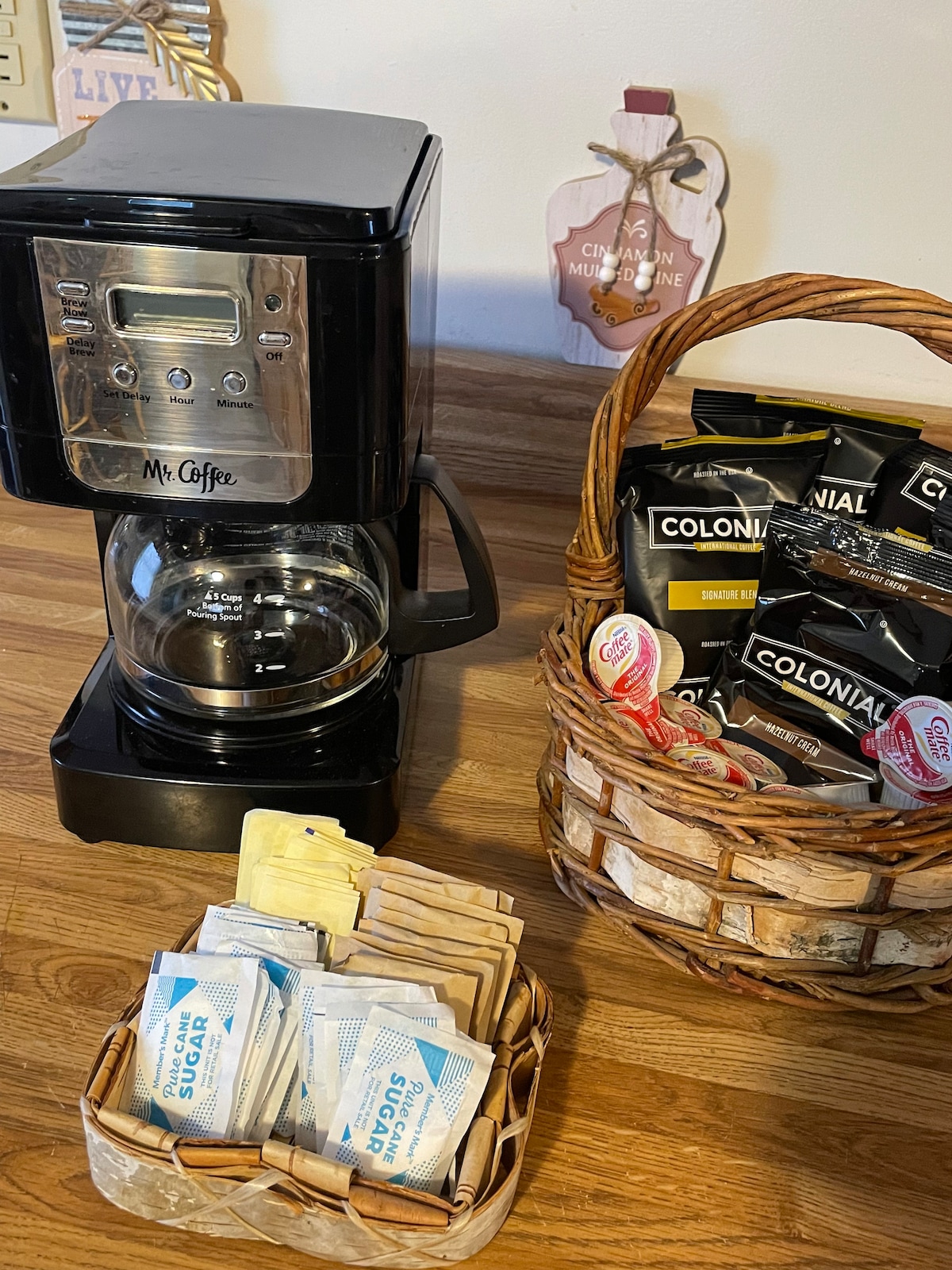 A coffee station is shown featuring a coffee maker next to a woven basket filled with coffee packets. A small container holds various sugar and creamer options, all arranged on a wooden countertop.