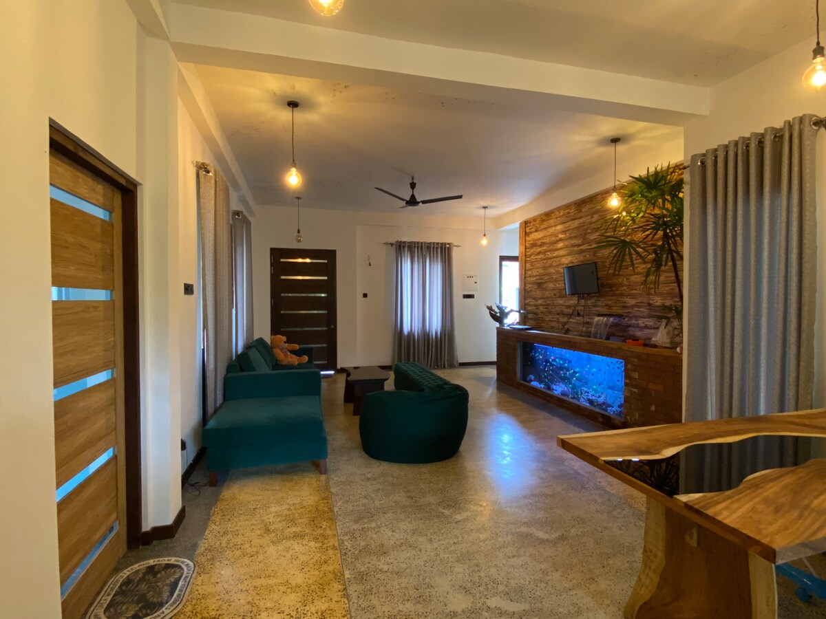 The living area features a combination of modern and natural elements. A teal sofa and bean bags are positioned around a wooden table. A wall-mounted television is visible above a console with an illuminated aquarium. Natural light enters through large windows adorned with drapes.