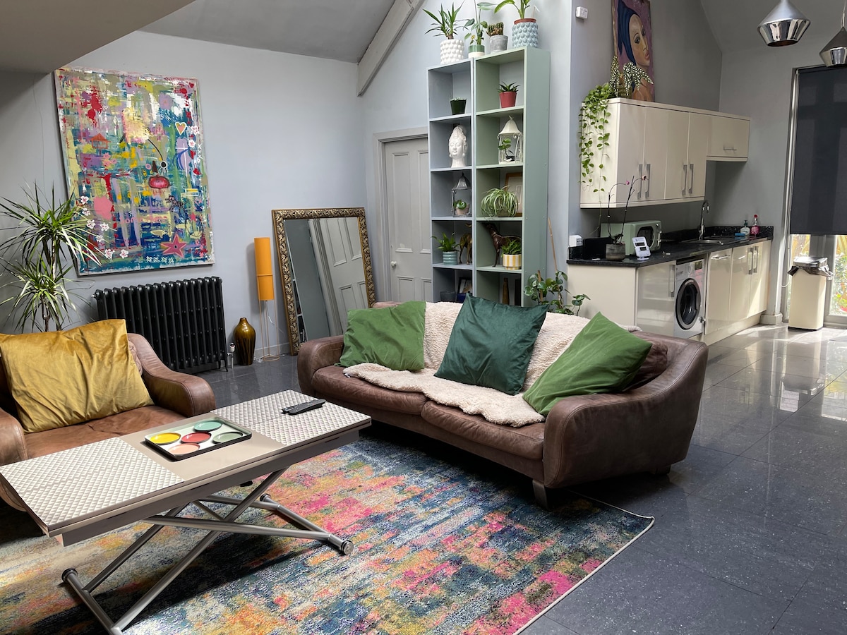 A spacious living area features a modern leather sofa adorned with green and golden cushions. A colorful abstract painting hangs on the wall above, complemented by a light rug. A fully equipped kitchen is visible in the background, with plants adding natural elements to the space.