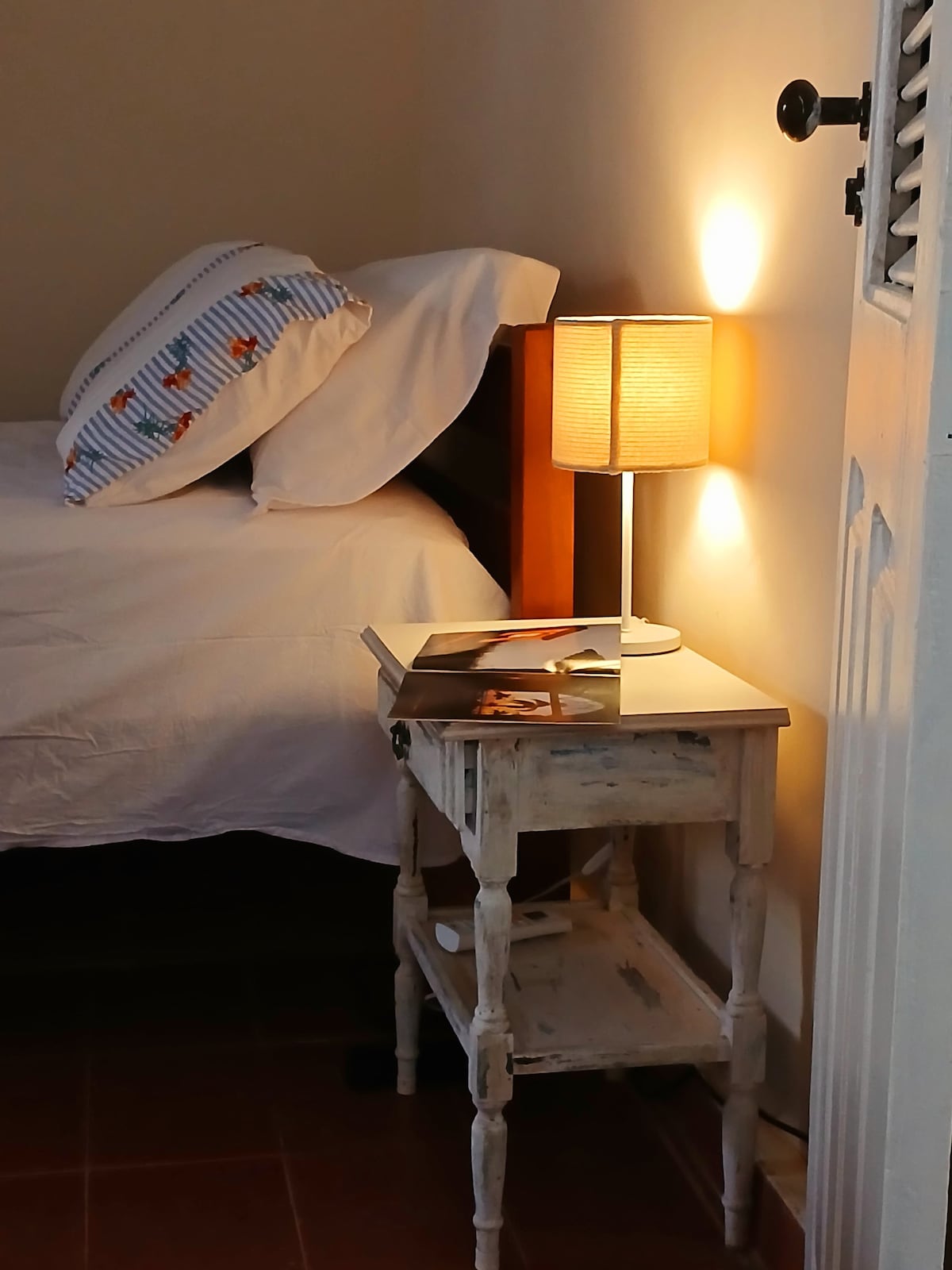 A neat bedroom corner features a single bed with patterned pillows and a warm light from a bedside lamp. A small, white nightstand stands beside the bed, holding reading materials and a decorative item, enhancing the functional yet cozy atmosphere of the space.