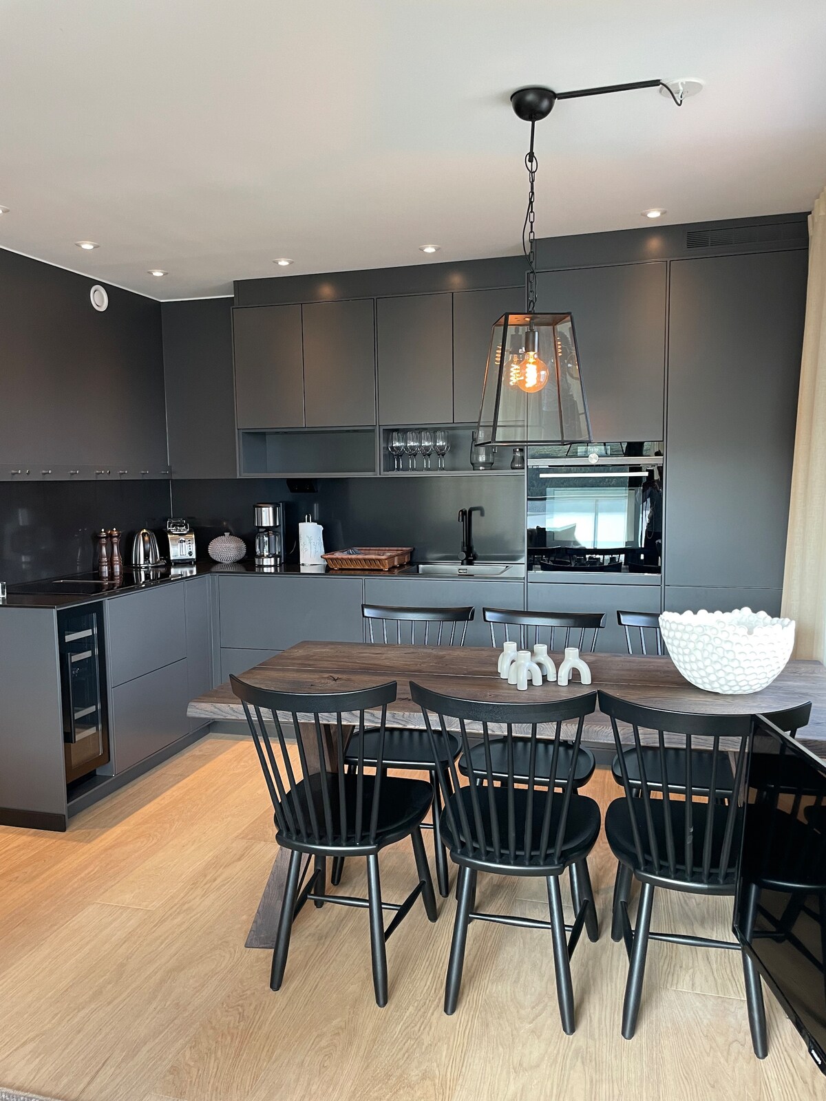 A modern kitchen is presented with sleek dark cabinetry and stainless steel appliances. A wooden dining table is surrounded by six black chairs. Soft lighting is provided by an industrial-style pendant fixture, enhancing the kitchen's contemporary feel. A bowl is placed at the center of the table.
