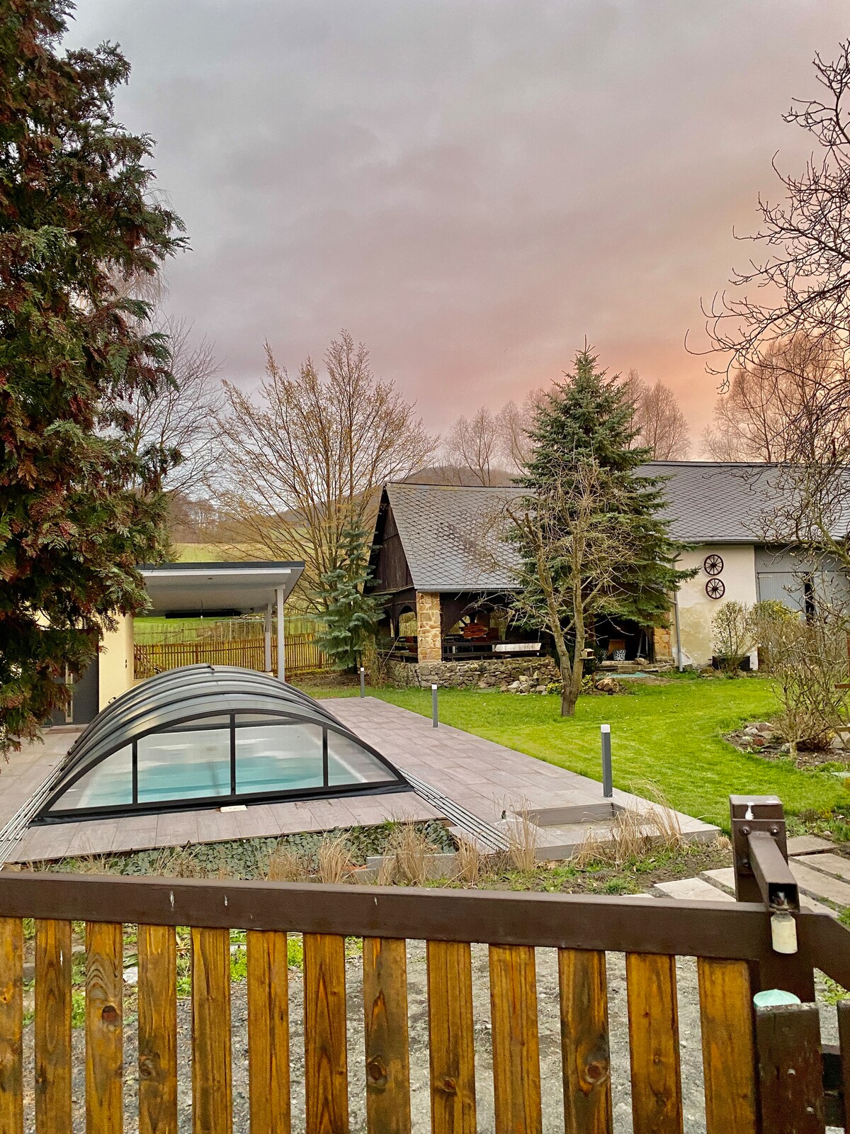 A serene outdoor view captures a landscaped garden with various trees and a covered swimming pool. A spacious patio is visible, accompanied by a rustic house featuring a sloped roof. The sky exhibits soft hues during sunset, enhancing the peaceful ambiance.