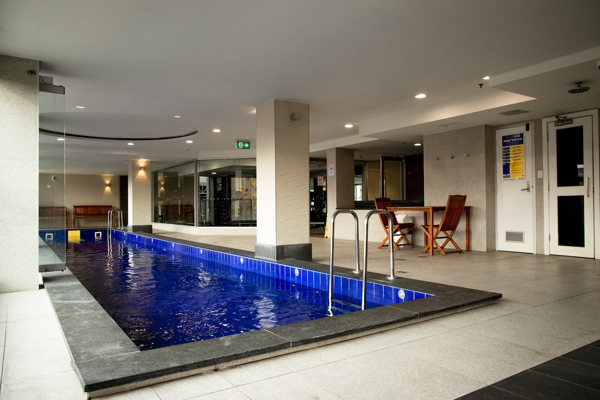A sleek indoor pool is surrounded by modern decor. The pool features blue tiles and is complemented by a set of wood outdoor furniture nearby. The area is well-lit with overhead lighting and offers a clean, spacious environment.