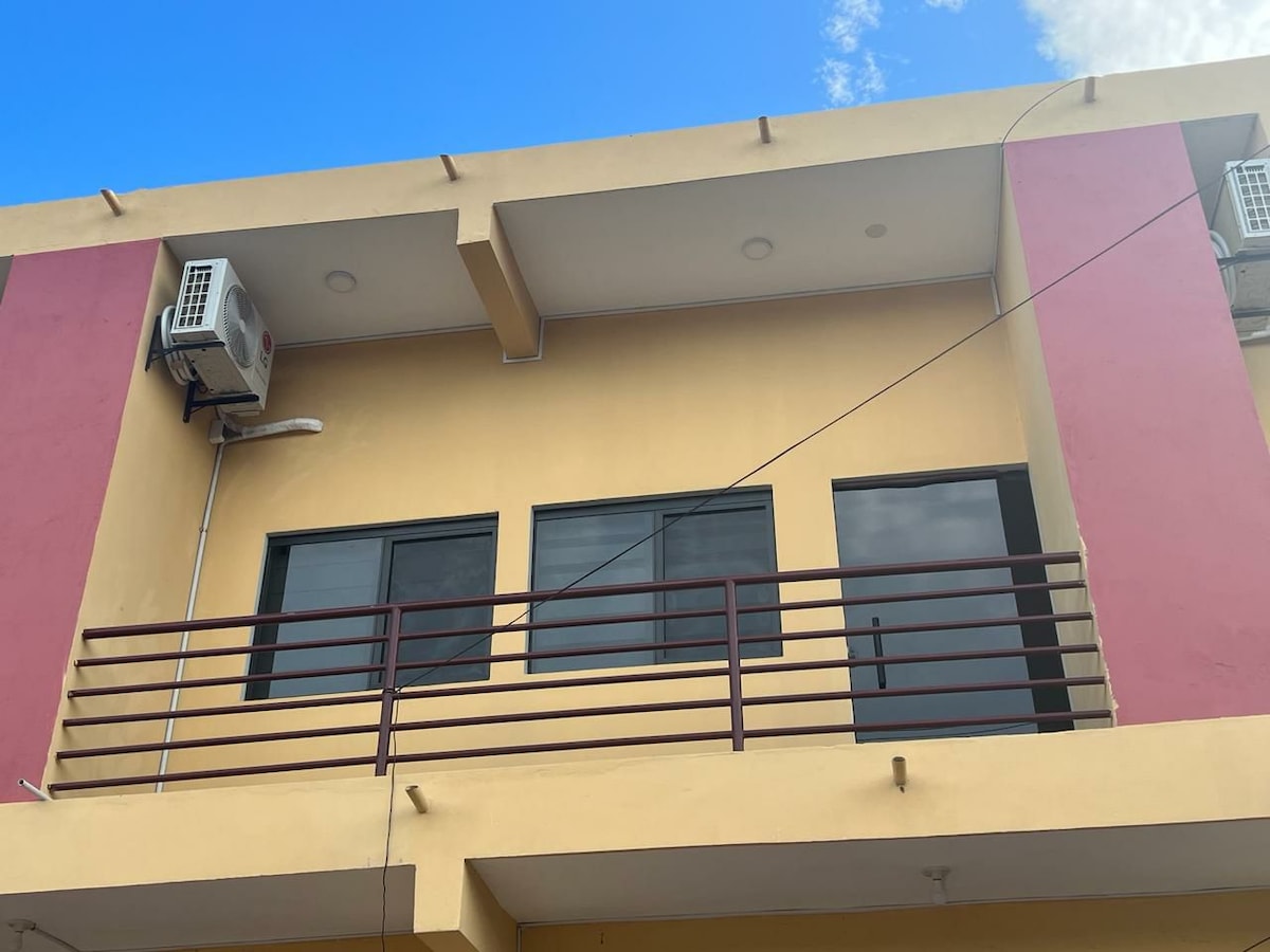 The exterior of a two-story building is visible, featuring large windows framed by a mix of yellow and pink walls. A balcony with a horizontal railing overlooks the ground level, and compact air conditioning units are mounted near the upper windows.
