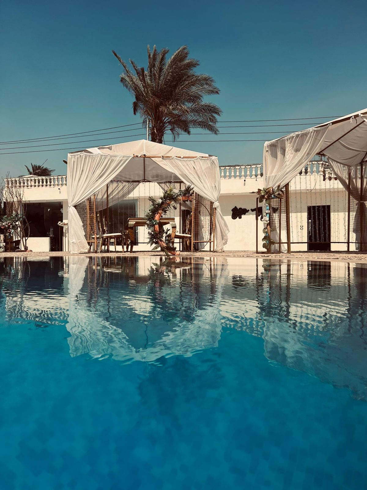 A clear blue swimming pool reflects the surroundings, including two elegant shaded cabanas with white drapes. In the background, a building with a simple design can be seen, accompanied by tropical palm trees swaying gently in the breeze.