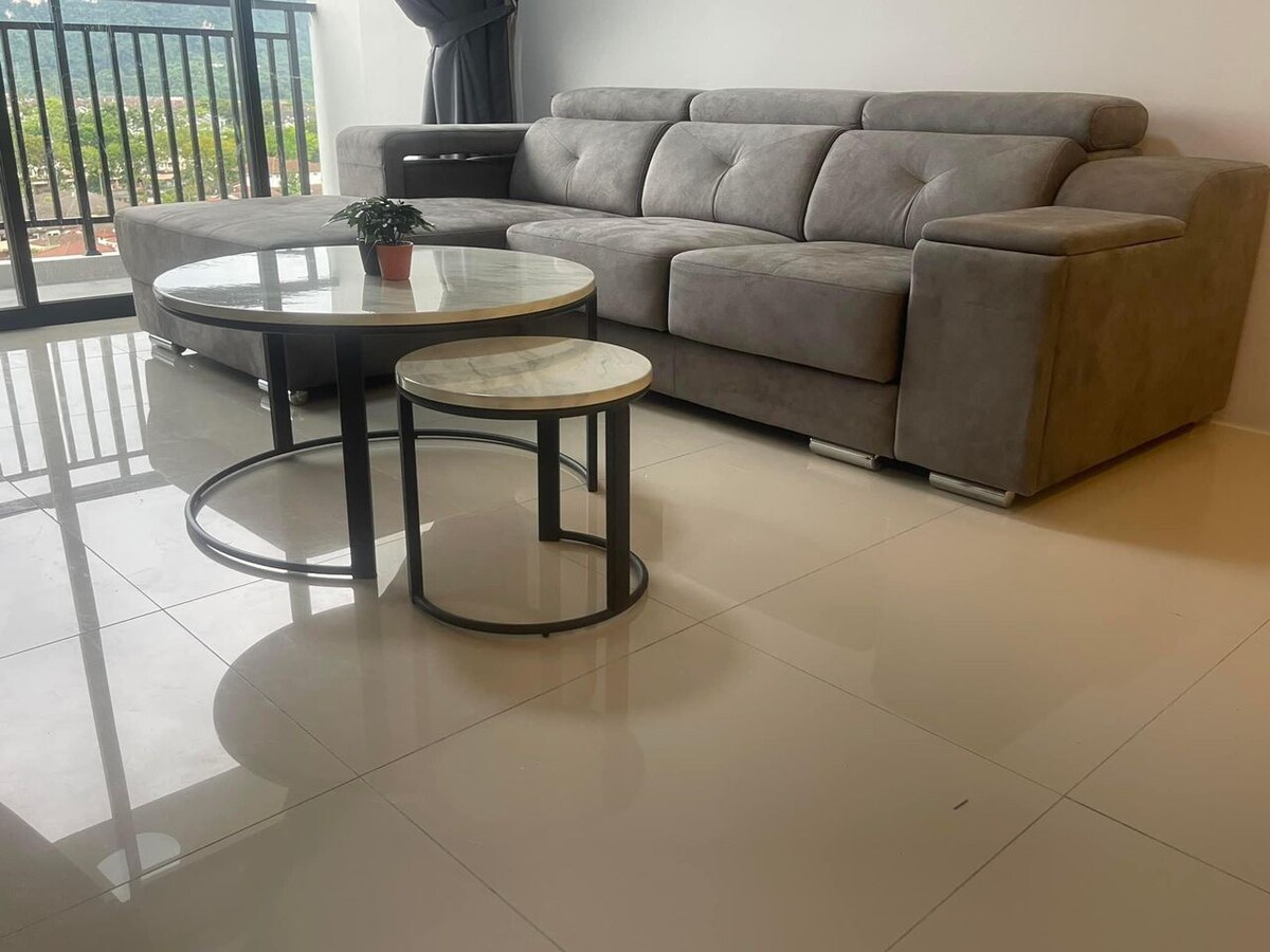 A modern living area is featured, showcasing a spacious gray sectional sofa beside two circular tables. The larger table has a white marble top and is complemented by a smaller, matching side table. Natural light streams in through the glass door, illuminating the tiled floor.