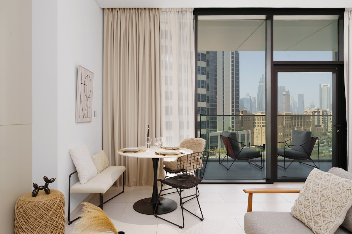 A modern dining area is presented with a round table and two stylish chairs, accompanied by a cozy bench. Floor-to-ceiling windows offer views of the city skyline, with sheer curtains allowing natural light to fill the space while maintaining privacy.
