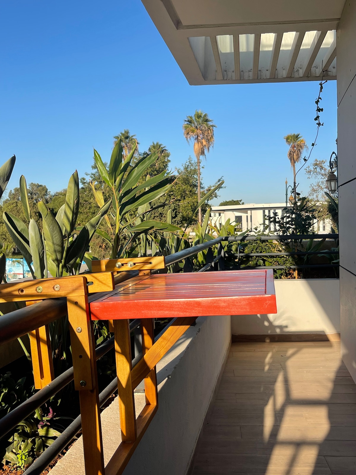 A balcony is shown with a red wooden table attached to the railing. Lush green plants are visible, alongside tall palm trees against a clear blue sky. The flooring is light-colored, contributing to a bright and airy atmosphere.