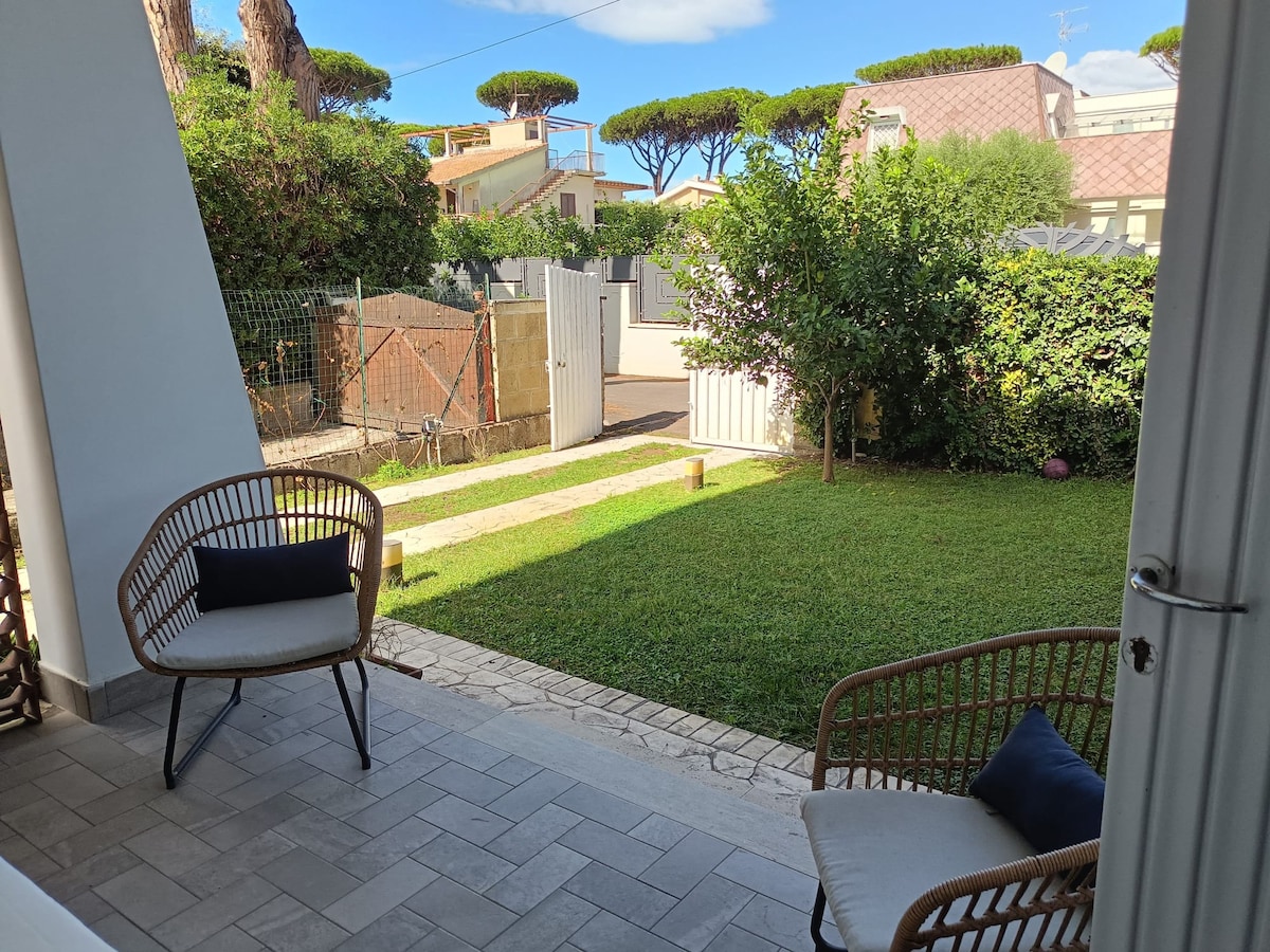 A serene outdoor space features two woven chairs positioned on a tiled patio, offering a view of the green lawn. Sunlight filters through the trees, creating a peaceful atmosphere. A pathway leads toward a gate, bordered by low shrubs.