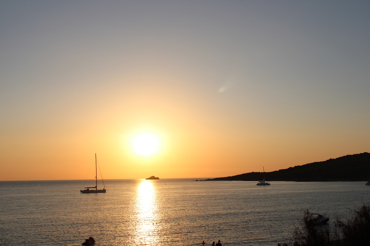 The image captures a serene sunset over calm waters, with the sun casting warm hues across the sky. Two boats are visible on the water, and a distant island can be seen along the horizon, creating a tranquil coastal scene.