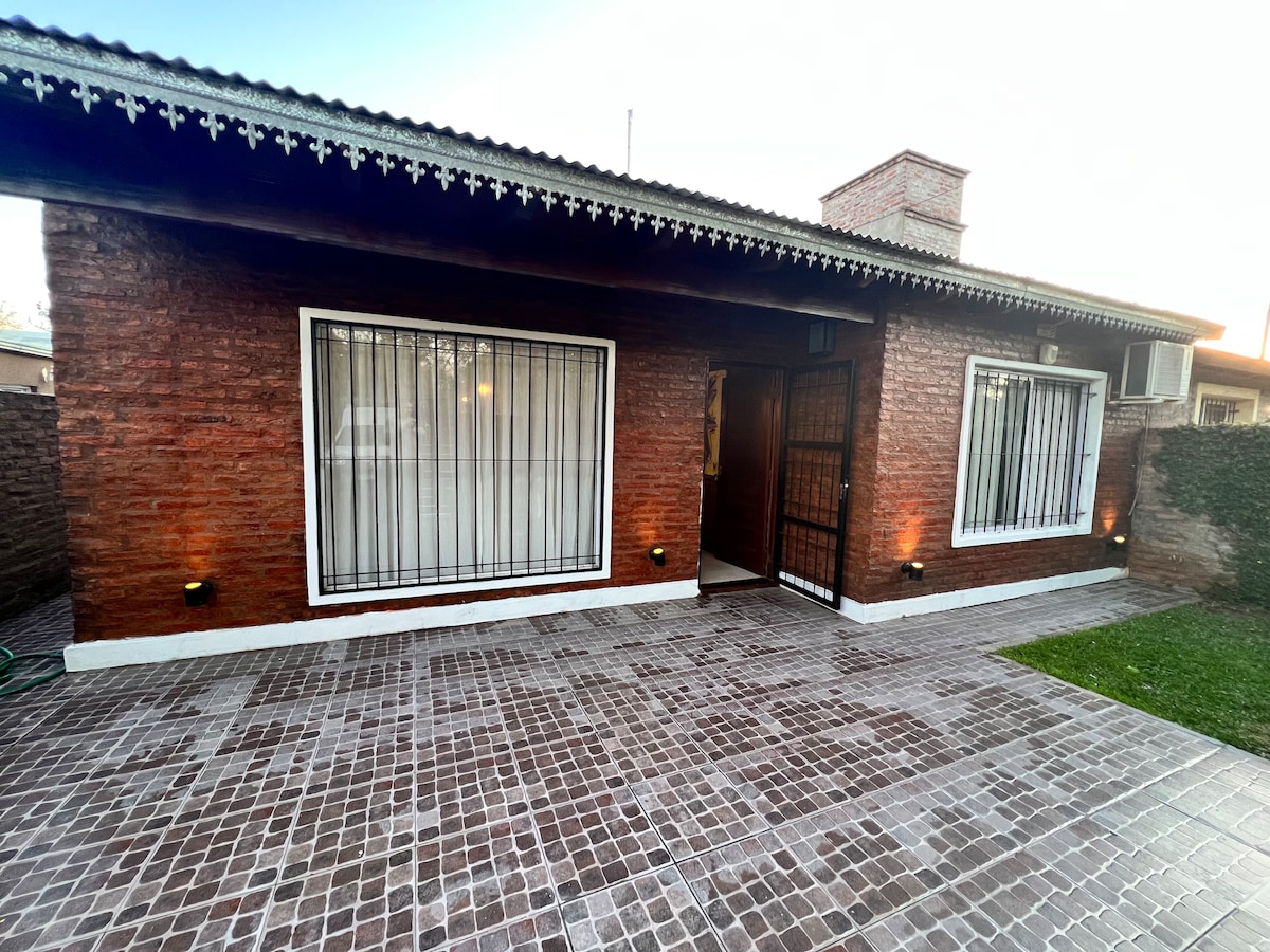 A brick exterior is showcased with two large windows featuring decorative metal grilles. The patio area is laid with square tiles, bordered by a green lawn and hedges, with outdoor lighting illuminating the entrance.