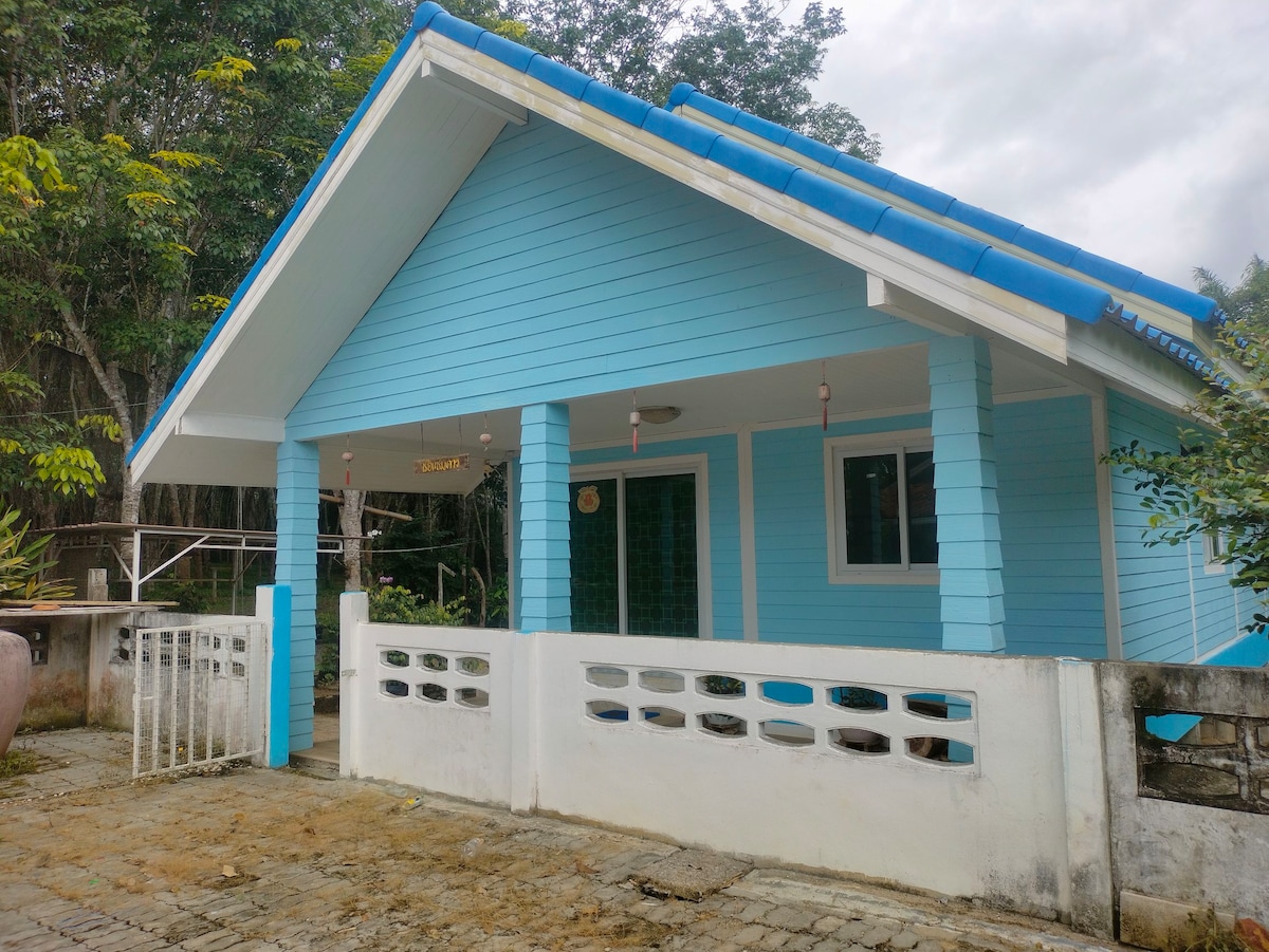 A charming light blue house features a sloped roof with blue tiles. The exterior is complemented by white accents, including a low wall at the front and a gated entrance. Lush greenery surrounds the property, creating a serene atmosphere.