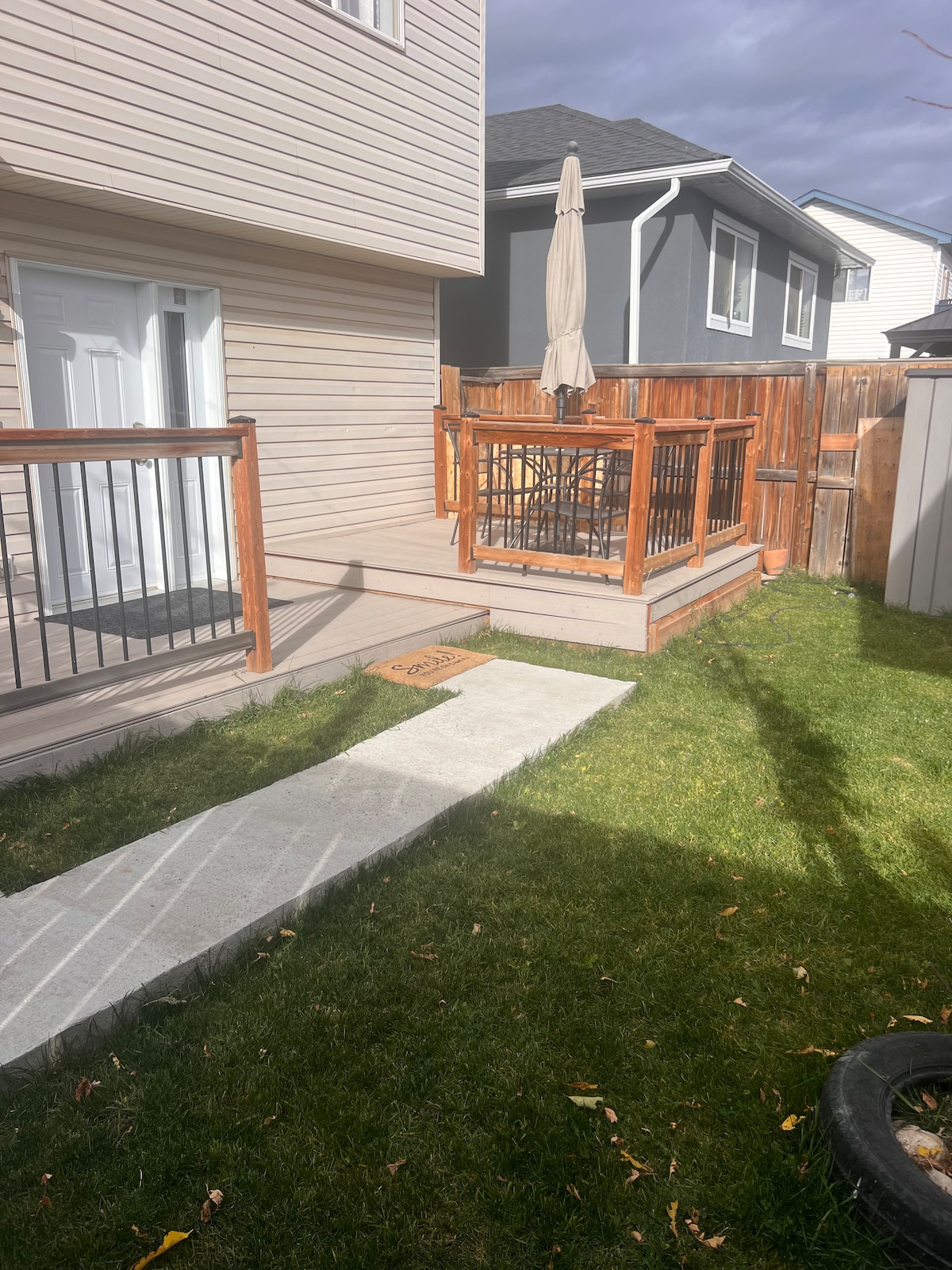 A well-maintained outdoor area is shown, featuring a concrete pathway leading to a wooden deck surrounded by a fenced yard. A patio umbrella is positioned over the seating area on the deck, while well-kept grass and a slight shadow indicate the time of day.