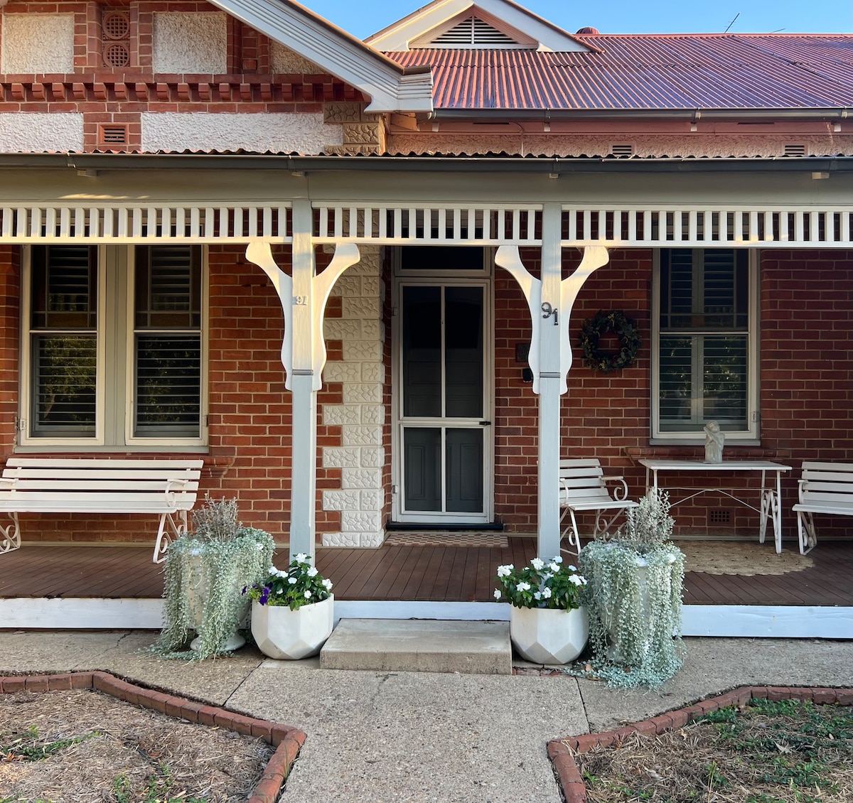 The cottage features a welcoming front porch with decorative white balconies and potted plants. Two white benches sit on the wooden deck, framed by a red brick facade. The entrance displays a screen door, complemented by a small table and chairs under the overhang.