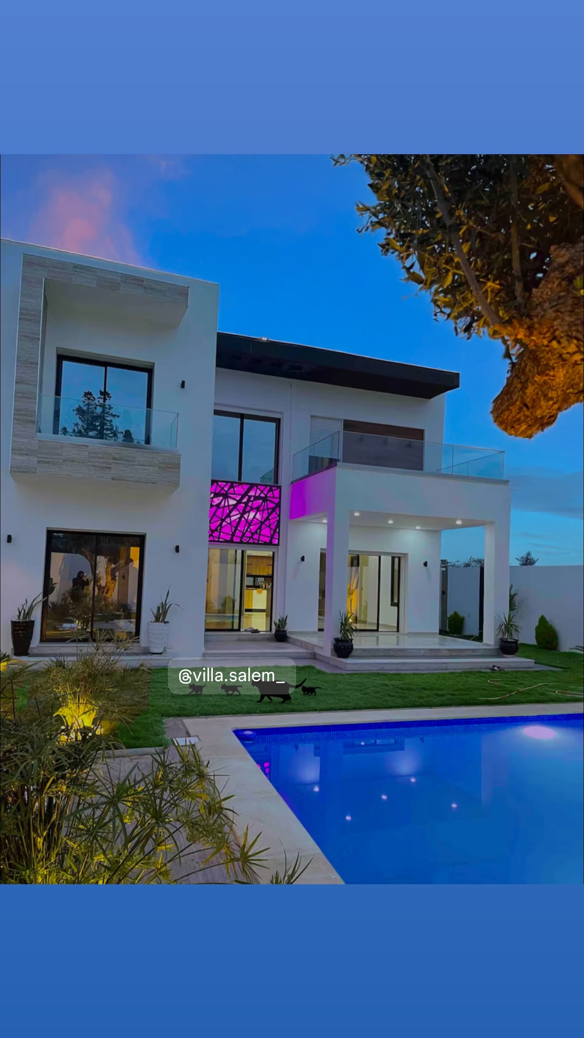 The exterior of a modern villa is presented at twilight, featuring a well-lit swimming pool and a neatly manicured lawn. Colorful lighting highlights the front facade, which includes large windows and a balcony. A tree adds a natural element to the scene.
