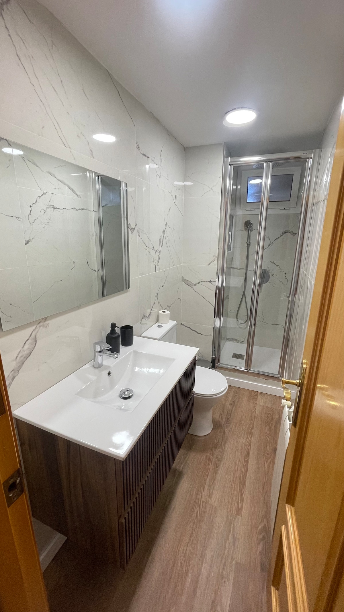 A modern bathroom is showcased, featuring marbled wall tiles and a sleek vanity with a sink and storage. A rectangular mirror is positioned above the vanity. A glass shower enclosure is visible, equipped with a handheld showerhead. Wood-style flooring adds warmth to the space.