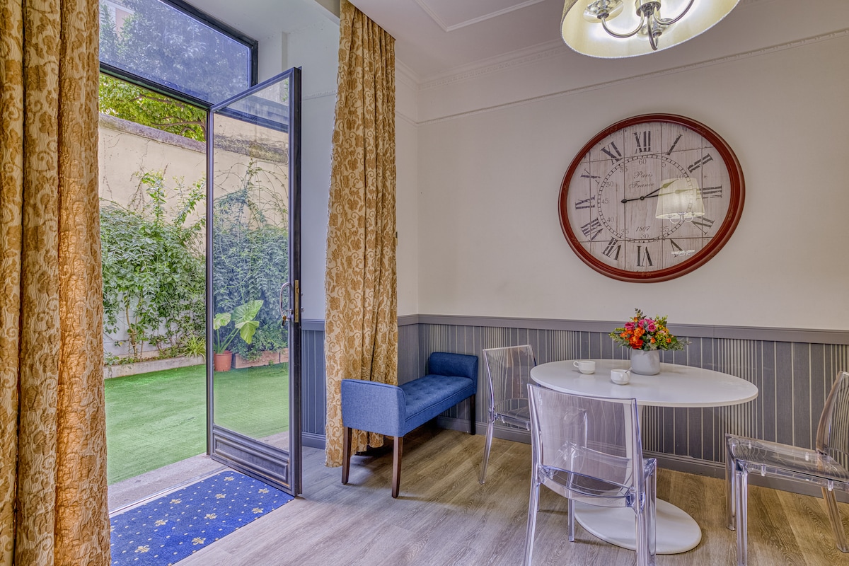Natural light fills a bright dining area, which features a round table surrounded by clear chairs. A blue upholstered bench adds a touch of color, while a large wall clock serves as a focal point. Floor-to-ceiling doors open to a small outdoor space with greenery.