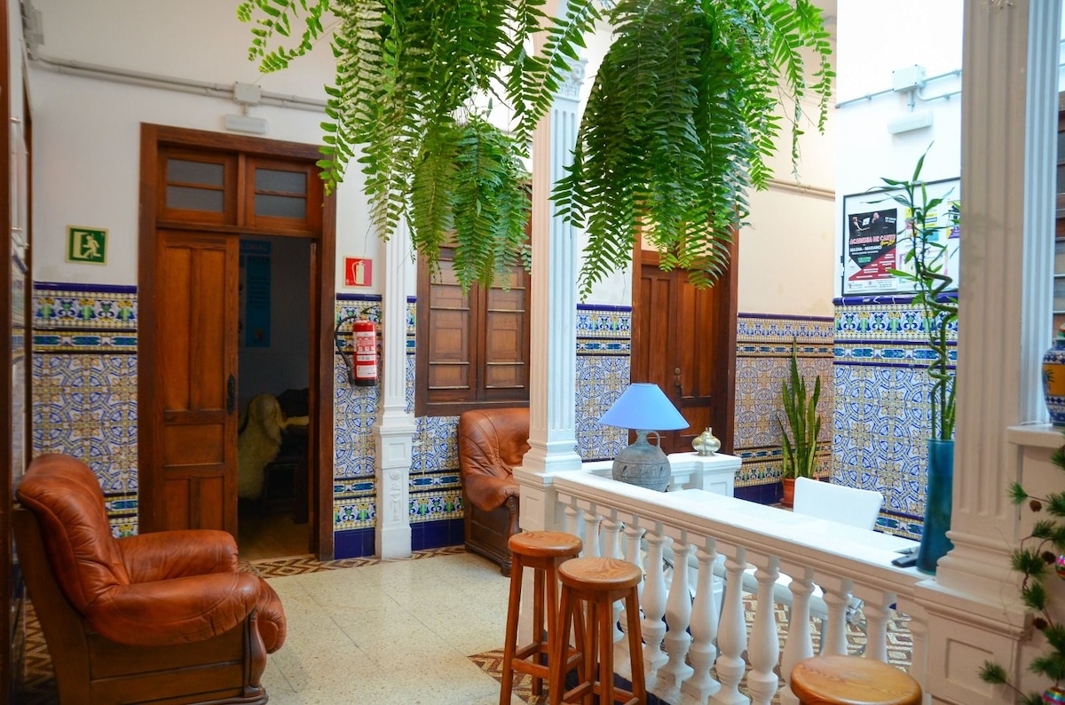 An inviting common area features leather seating and a light blue lamp on a white table. Lush green plants hang from the ceiling, adding a fresh touch. The tiled walls are adorned with intricate patterns, contributing to the space's cultural character.