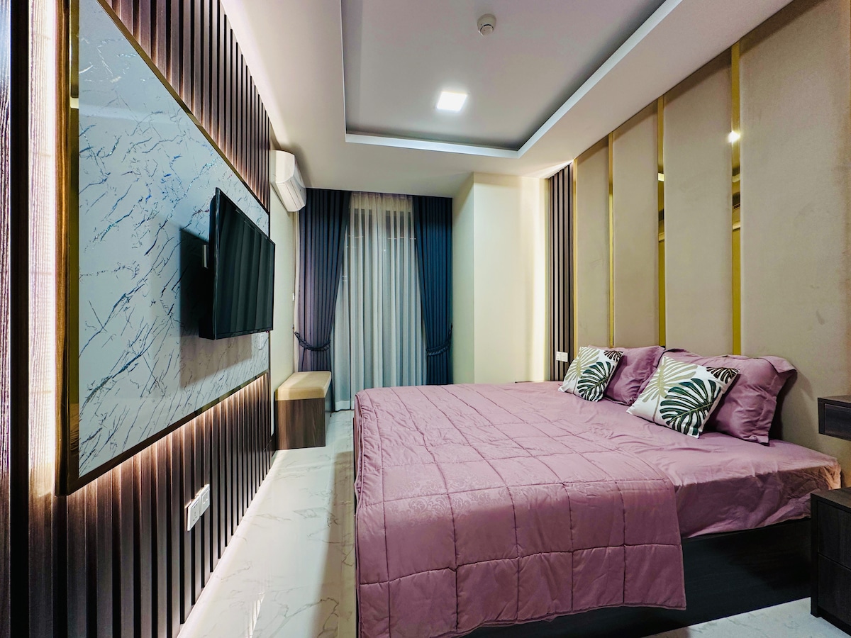 A modern studio room features a spacious bed dressed in a soft pink coverlet. The wall-mounted television and elegant decor enhance the minimalist design. Large windows with sheer curtains allow natural light to fill the room, complemented by warm wood accents and stylish wall finishes.