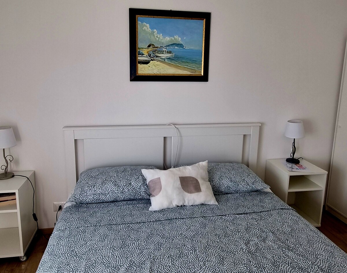 A bright bedroom features a spacious bed adorned with a patterned blue coverlet. Simple bedside tables with lamps sit on either side. Above the bed, a framed painting of a coastal scene adds a touch of color to the neutral walls.