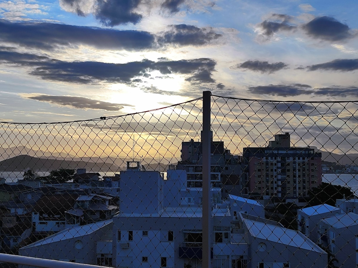 A view of the horizon showcases a colorful sunset over the city, with clouds subtly reflecting the fading light. A protective mesh fence adds a layer of safety, framing the landscape that includes buildings and distant hills under a dynamic sky.