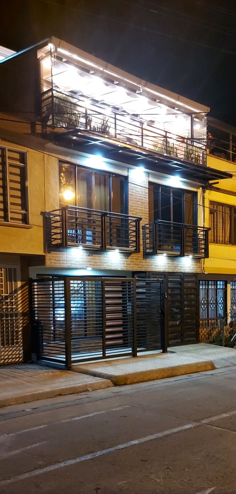 A two-story building is illuminated at night, showcasing a modern façade with large glass doors and balconies. The ground level features a gated entrance with sleek metal detailing, while the upper level is highlighted by warm lighting beneath a pergola.