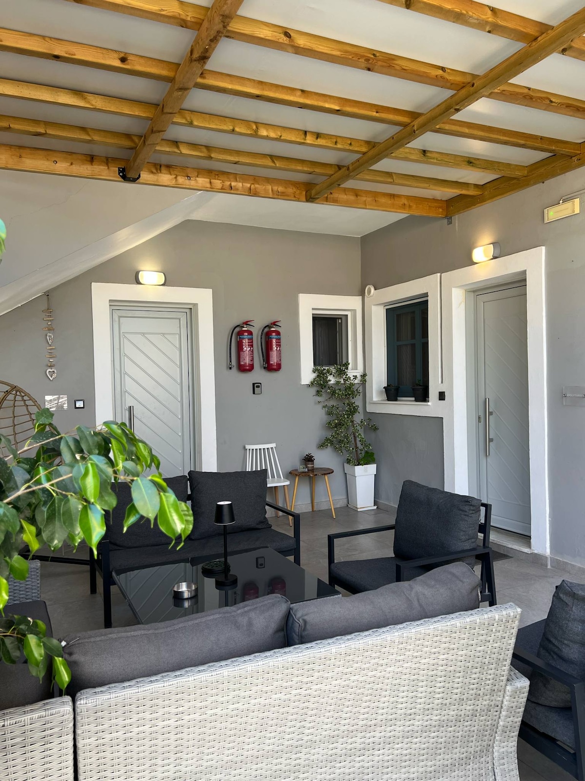 A shared outdoor seating area is presented, featuring a dark glass table surrounded by comfortable gray chairs. The textured ceiling with wooden beams and bright lights enhances the ambiance. Two separate entrances with gray doors are visible, alongside plants in decorative pots.
