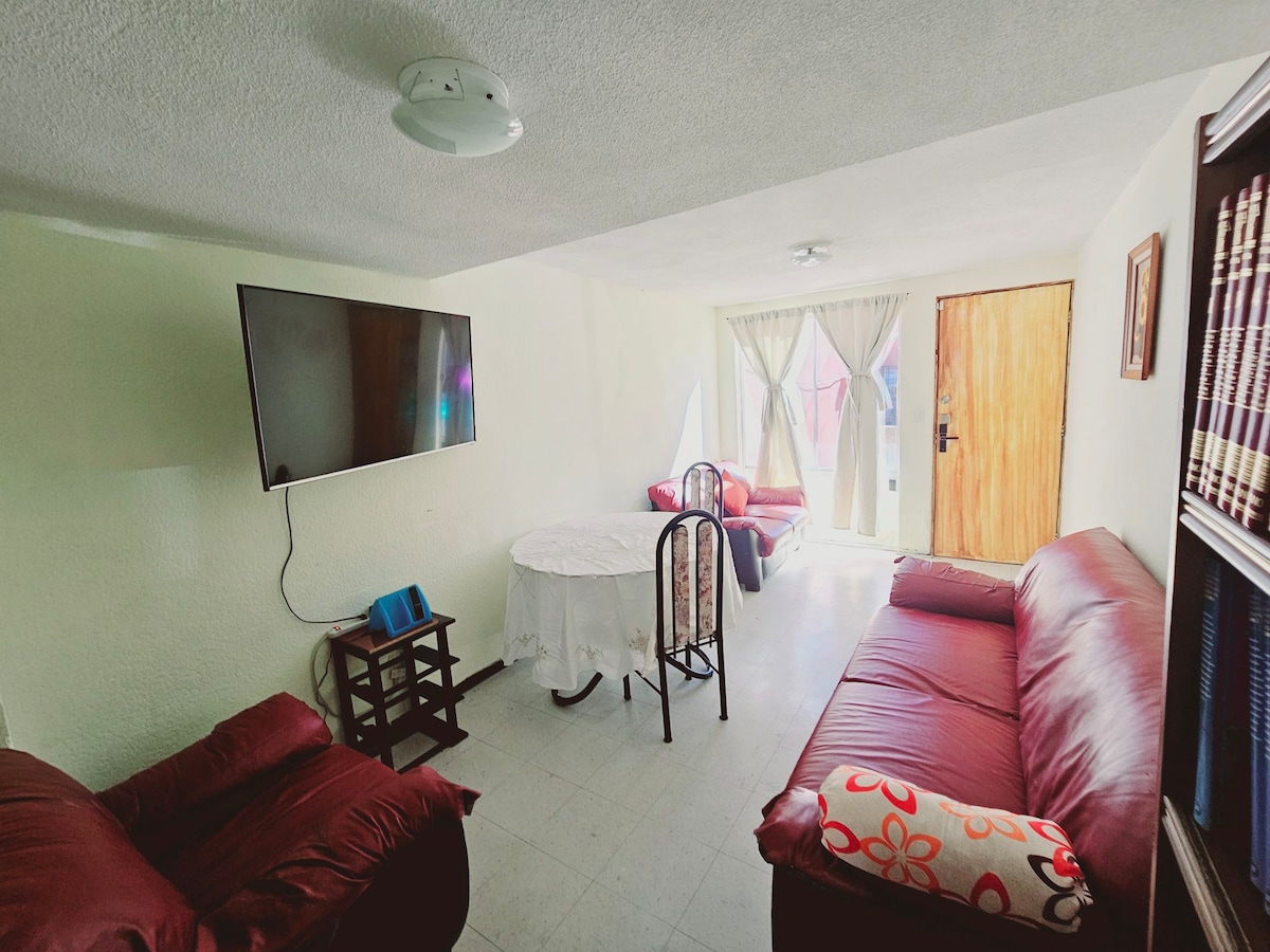 The living room features a large flat-screen TV mounted on the wall and two red sofas positioned for comfort. A round dining table with chairs is set near the window, which is adorned with sheer curtains, allowing natural light to fill the space.