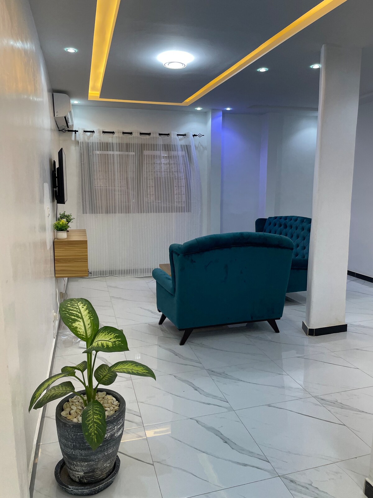 A modern living area is presented with two teal armchairs, complemented by a small wooden table. Natural light filters through sheer curtains. A decorative plant in a textured pot adds a touch of greenery to the polished marble floor.