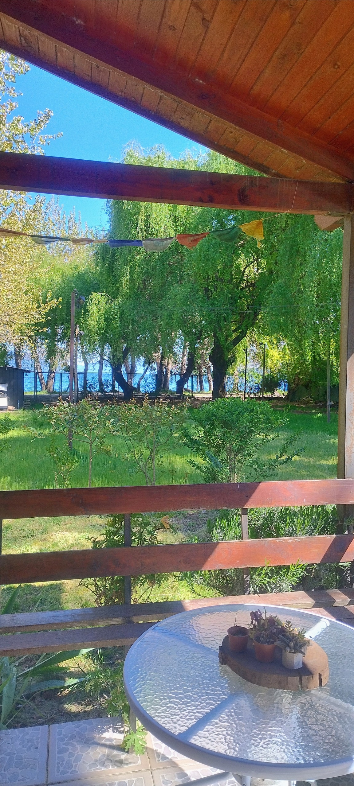 A serene outdoor view is captured from a wooden deck overlooking lush greenery and a lake. A round glass table is visible in the foreground, adorned with decorative elements. Willows sway gently in the breeze, enhancing the tranquil setting.