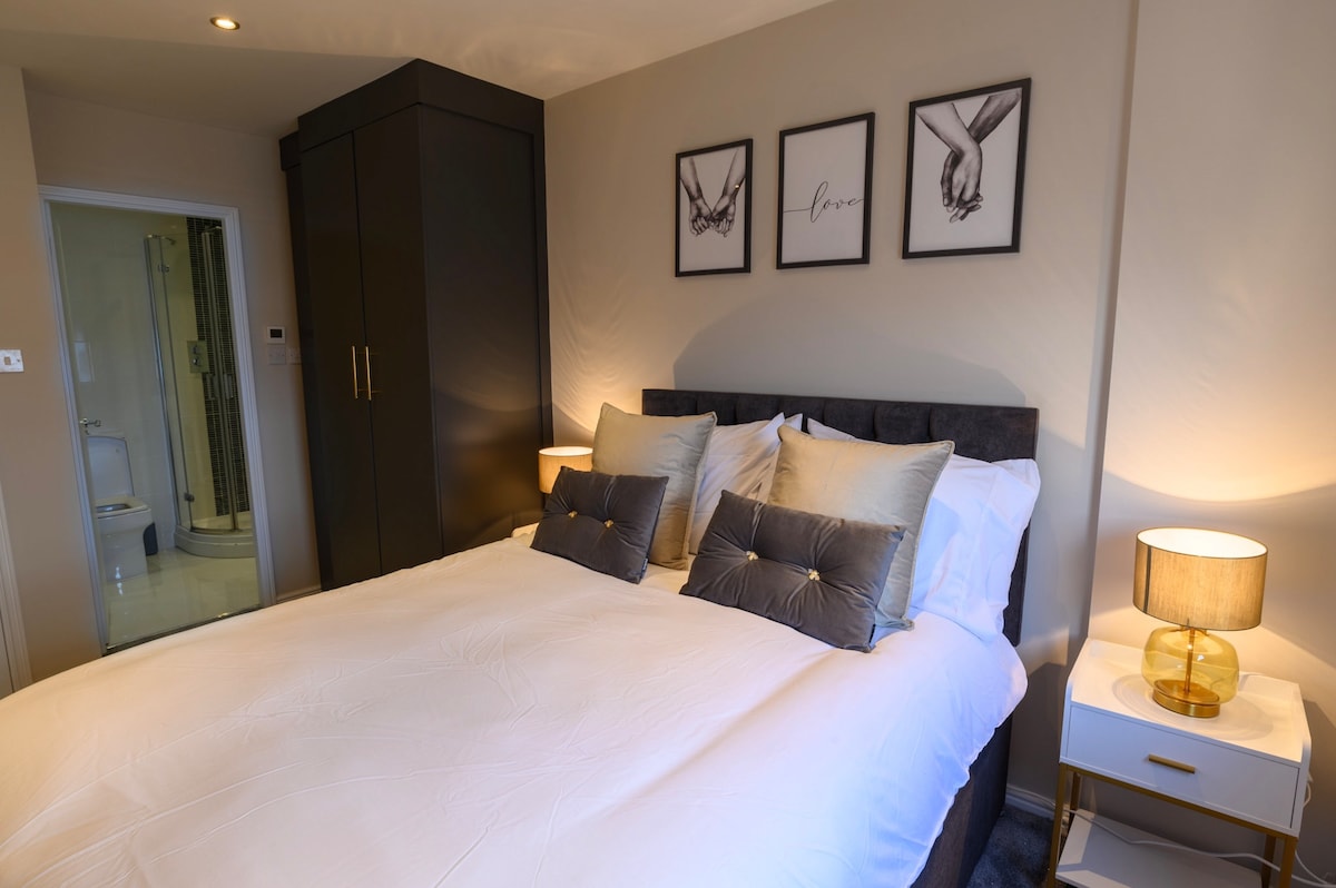 A well-appointed bedroom features a spacious bed with plush pillows and decorative cushions. Framed artwork is displayed above the bed, complementing the contemporary décor. A warm bedside lamp casts a soft glow, while an en-suite bathroom can be seen in the background.