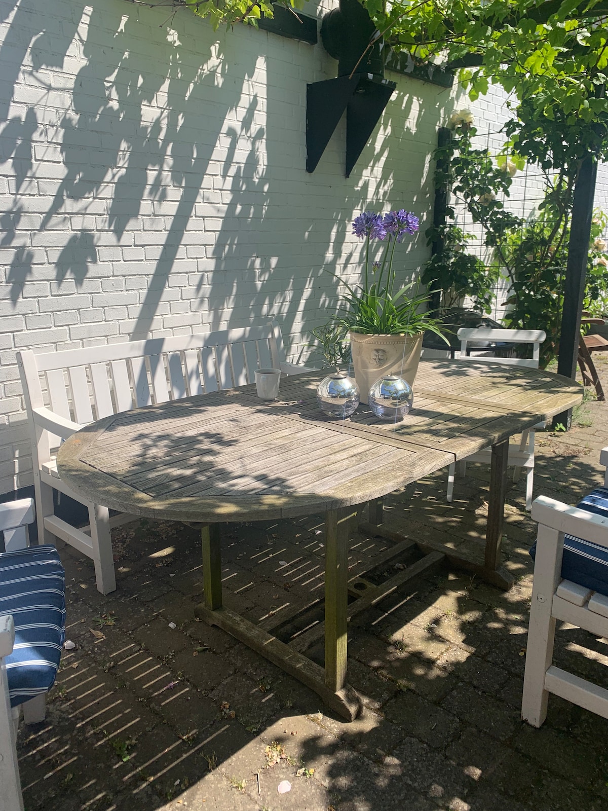 A wooden dining table is surrounded by white chairs in a shaded garden area. There are decorative plants and flowers placed on the table, with sunlight creating playful shadows on the brick wall and paving below.