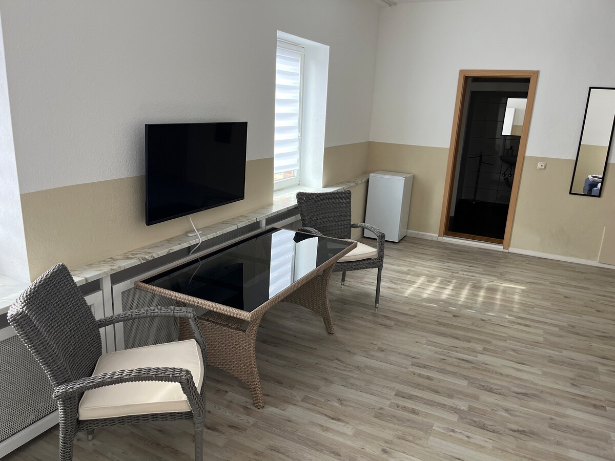 A spacious seating area contains a glass-topped coffee table with wicker chairs. A flat-screen television is mounted on the wall. Natural light streams in through a window with horizontal blinds, while a small refrigerator is visible in the corner.