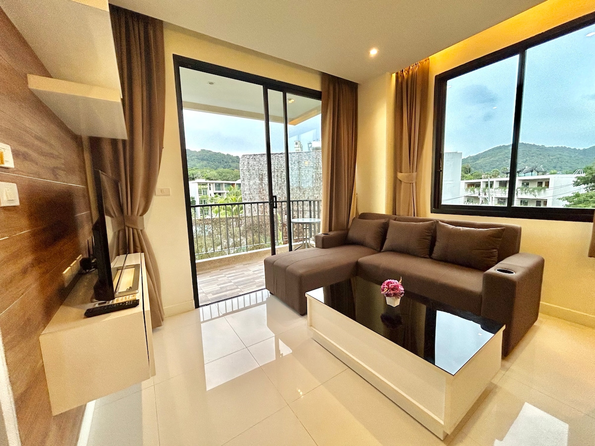 A living area is presented, featuring a modern sectional sofa and a coffee table with a small decorative piece. Floor-to-ceiling glass doors allow natural light to fill the space, while scenic views of the surrounding landscape are available from the balcony.