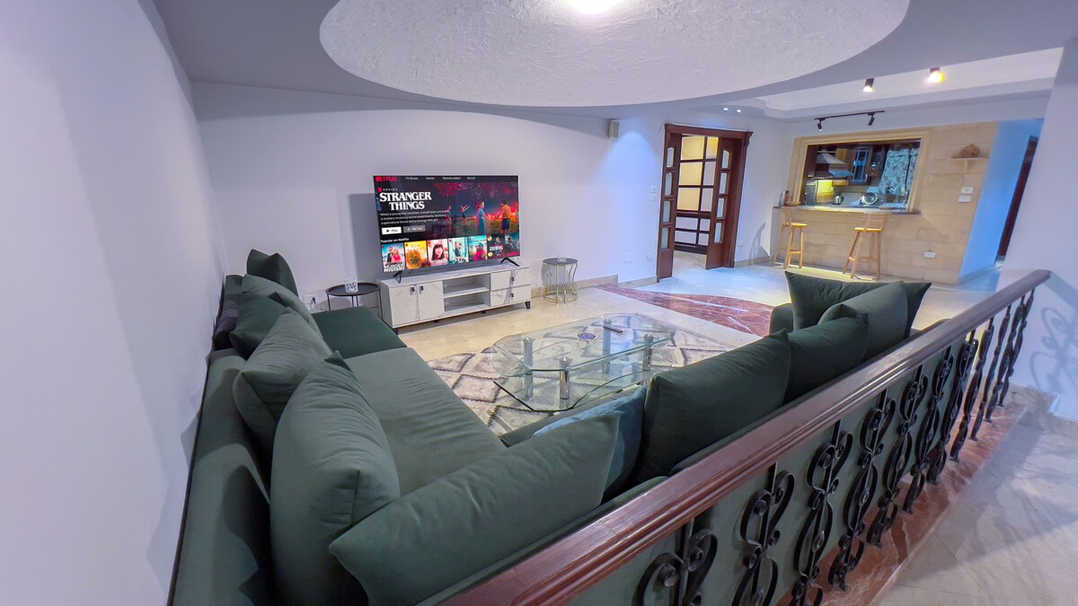 A spacious living area features a large sectional sofa in a muted green hue, oriented towards a flat-screen television mounted on a sleek entertainment unit. A glass coffee table complements the space, and an open kitchen area can be seen in the background, enhancing the inviting layout.