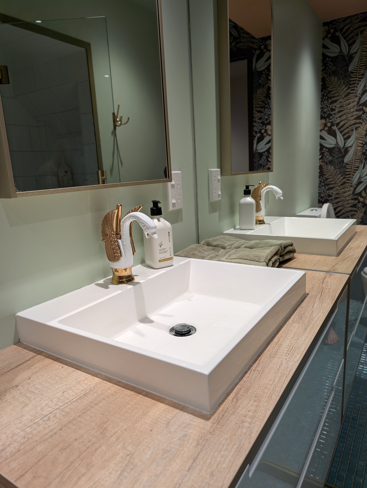 A contemporary bathroom features a spacious white sink set on a light wooden countertop. Two mirrors framed in gold are mounted above, and a decorative wall with leaf patterns adds a touch of nature. A roll of soft towels is neatly placed beside the sink.