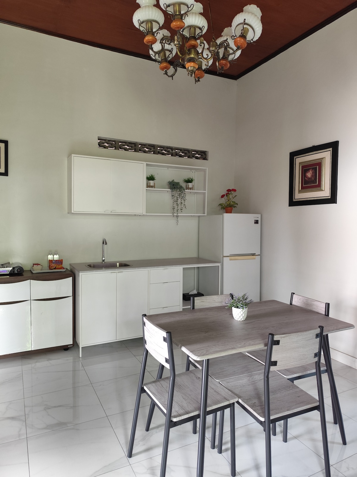 A dining area is showcased with a wooden table surrounded by four chairs. In the background, a modern kitchen is visible, equipped with cabinets, a sink, and a refrigerator. A decorative chandelier hangs above, and light-colored walls enhance the bright and airy atmosphere.