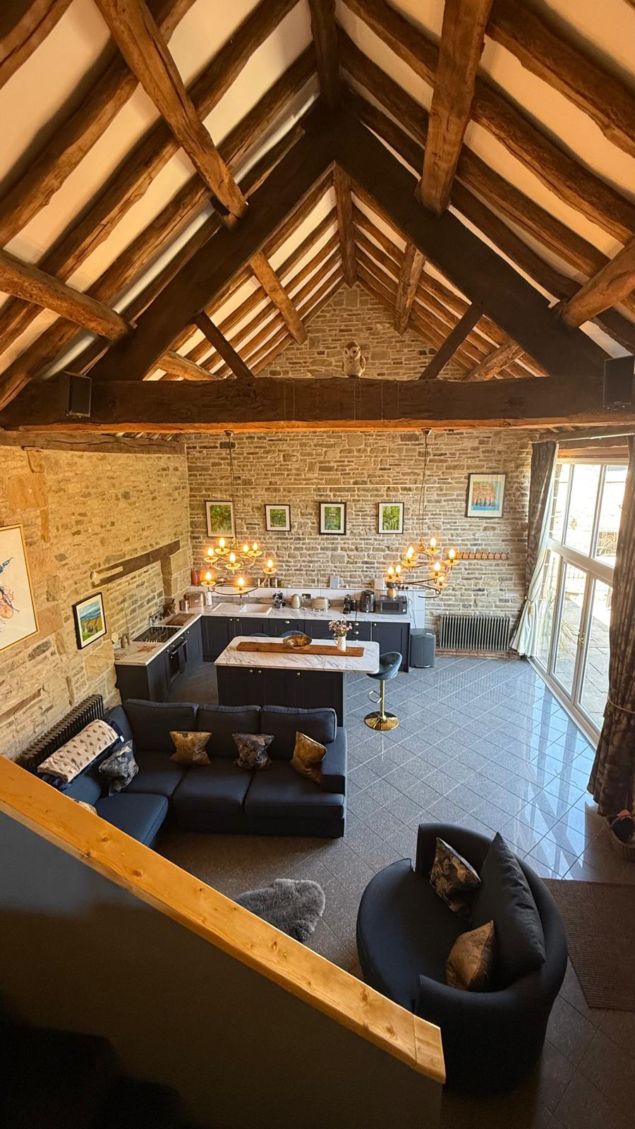 The image showcases the spacious living area of the restored barn, with high vaulted ceilings and exposed beams. An open kitchen features modern appliances, while comfortable seating is arranged to facilitate conversation. Large glass doors invite natural light and provide a view of the outdoor space.