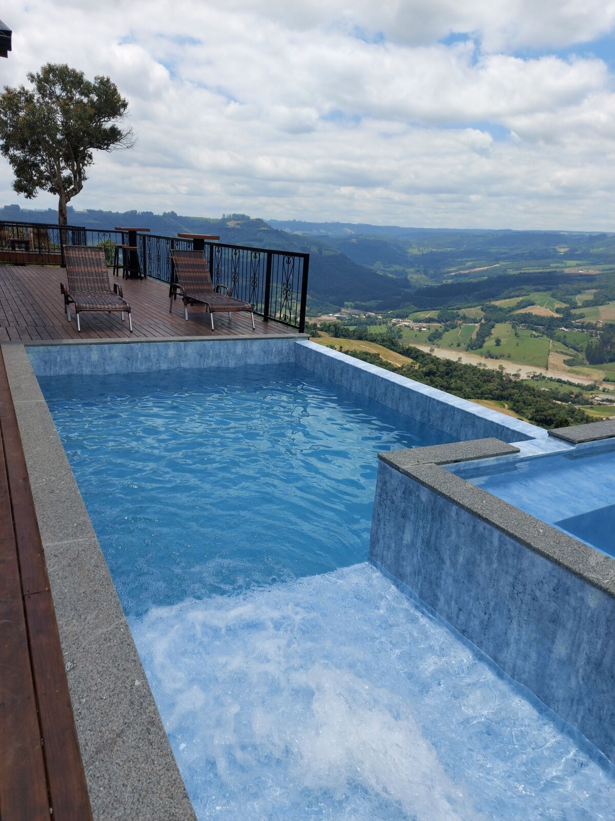 A serene infinity pool is visible, overlooking rolling green hills and a vast sky. Wooden lounge chairs are positioned on the deck, inviting relaxation. The pool features a smooth edge, creating a seamless view of the landscape beyond.