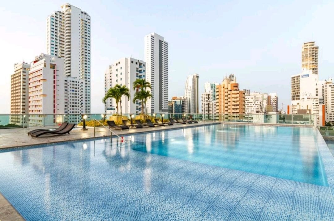 A rooftop swimming pool features shimmering turquoise tiles, surrounded by lounge chairs with yellow cushions. The skyline of Cartagena is visible in the background, showcasing several tall buildings against a soft sky.