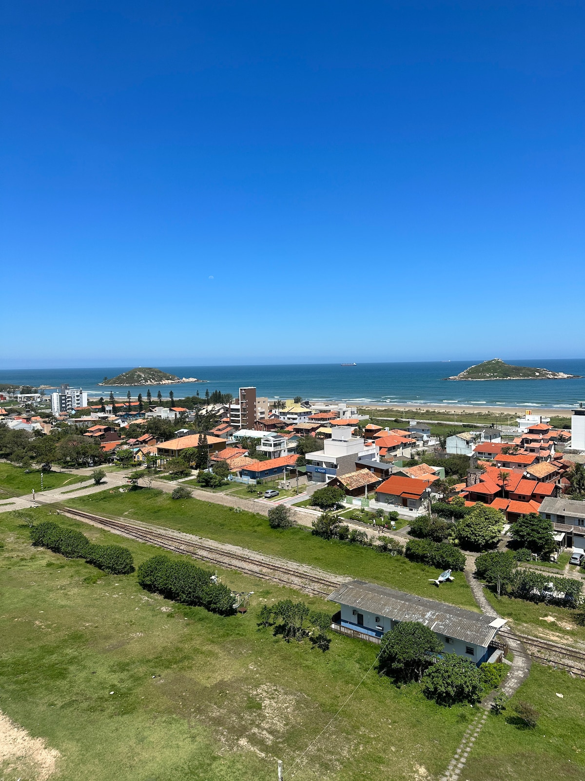 A panoramic view showcases a coastal landscape featuring the ocean and two small islands in the distance. The foreground includes a vibrant mix of buildings and homes, complemented by lush greenery and a railway line. The clear blue sky enhances the overall scene.