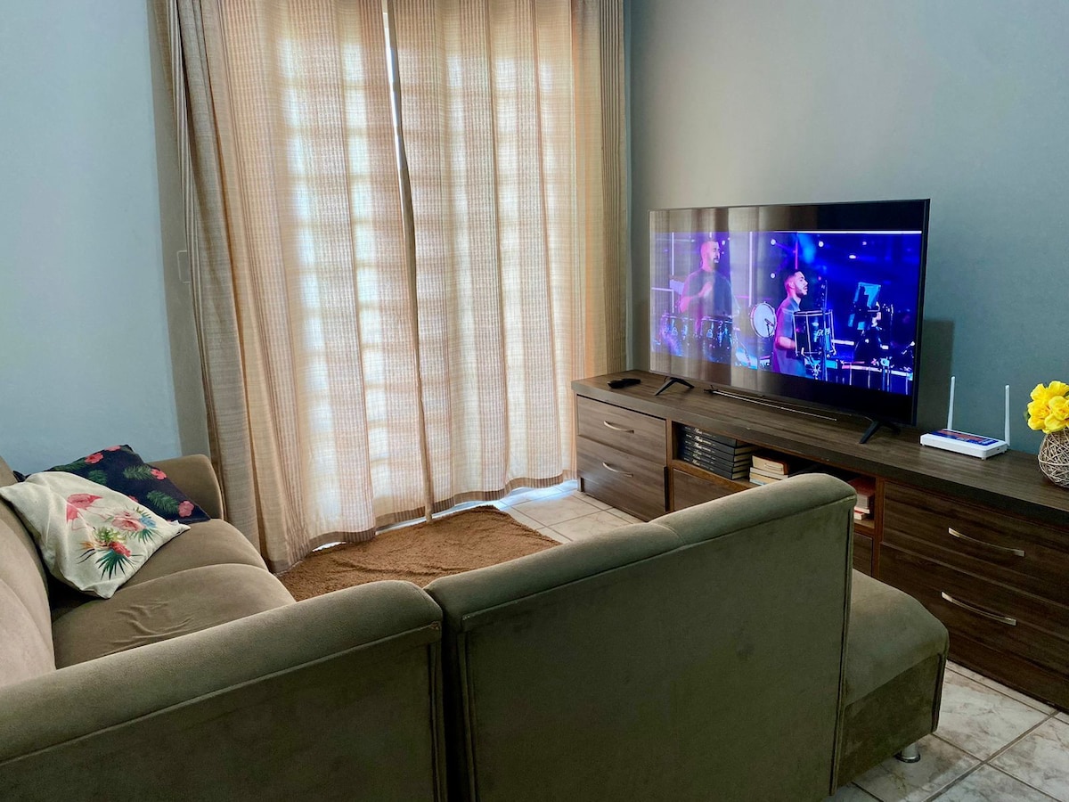 A comfortable seating area is featured, with a plush sectional sofa facing a flat-screen television. Light beige curtains diffuse natural light, and a small wooden TV stand is positioned beneath the screen. A soft area rug lies on the tiled floor, adding warmth to the space.