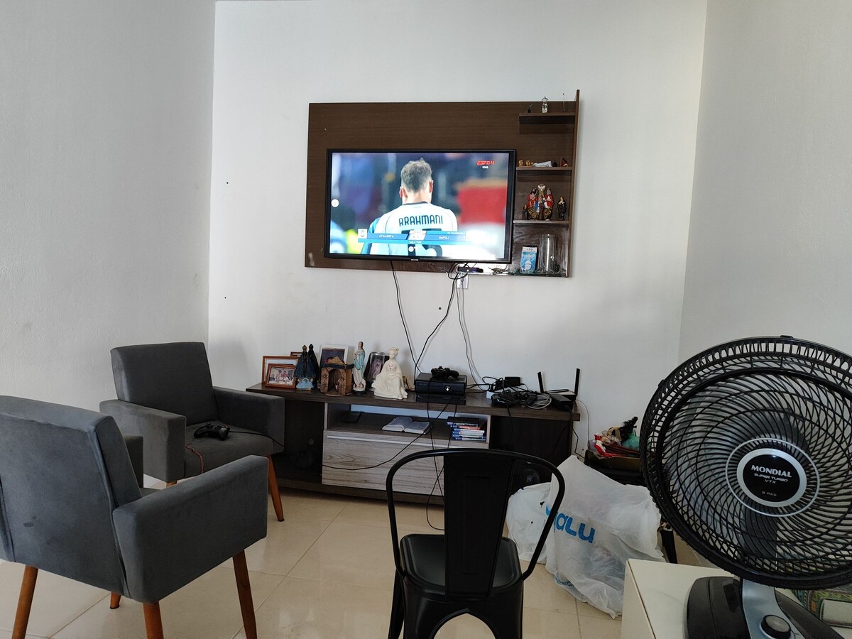 A relaxed living area is depicted with two grey armchairs facing a wall-mounted television. A wooden shelf holds framed photos and decorative items. A fan stands in the corner, and various items are arranged on the floor, contributing to the casual ambiance.