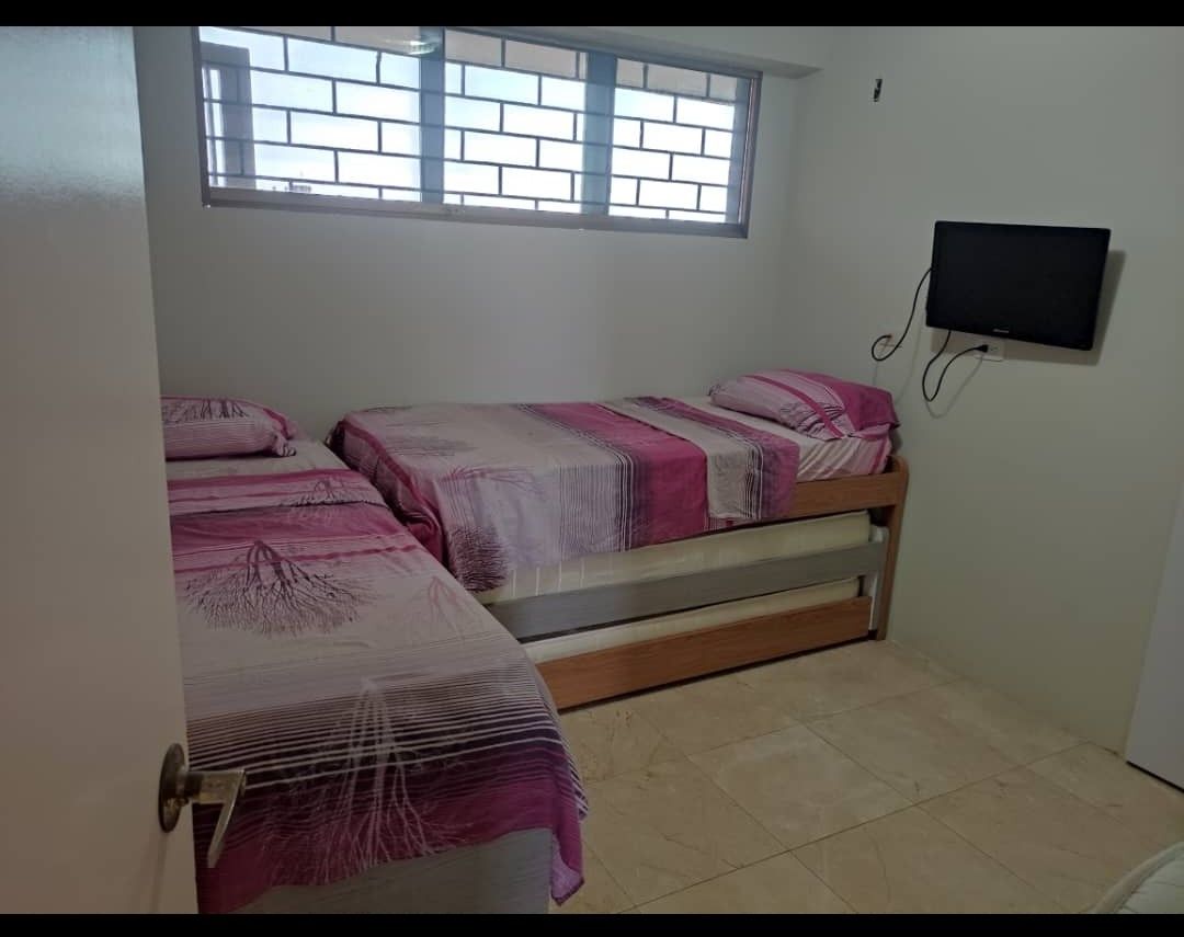 The room features two twin beds, each dressed with pink and white patterned linens. A window with a modern grid design allows natural light to fill the space. A flat-screen television is mounted on the wall opposite the beds, enhancing the room's functionality.