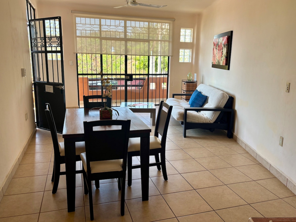 A bright living space is visible, featuring a dining table with four chairs and a comfortable sofa. Large windows allow natural light to fill the room, while a simple piece of wall art adds a touch of color. The entrance door opens towards a shared patio.