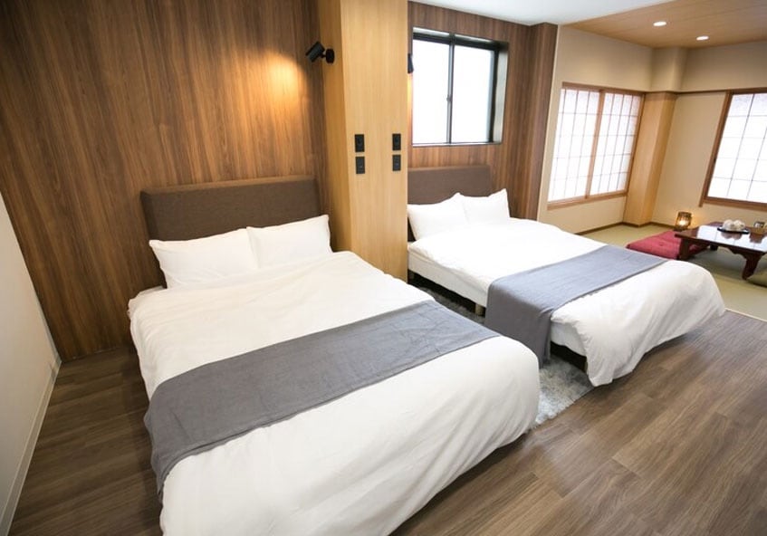 A serene bedroom features two spacious beds dressed in crisp white linens, complemented by light grey throws. Natural light floods the room through a large window, illuminating the wooden accent walls and soft carpet beneath. Sliding paper doors add a traditional touch to the overall design.