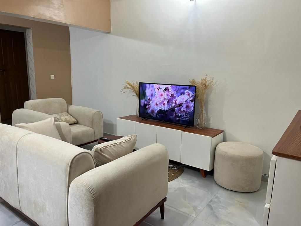 A comfortable living area is presented, featuring a soft beige sofa set arranged around a stylish coffee table. A television sits atop a low, white sideboard, while decorative plants add a touch of nature to the space. Light reflects off the smooth, tiled floor.