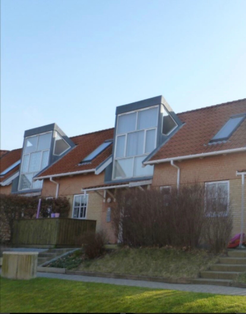 A two-story townhouse is depicted, featuring a sloped roof with large glass windows positioned at the front. The facade is made of light brick, and the yard displays a grassy area with steps leading up to the entrance. Bushes provide landscaping along the pathway.